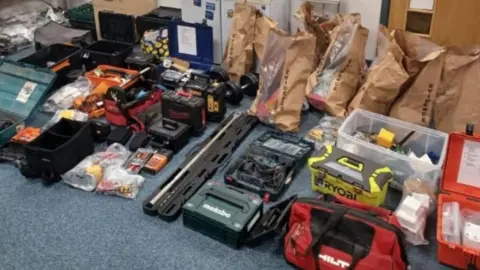 Boxes and bags of power tools and other tools lie on a carpeted floor in a room. 