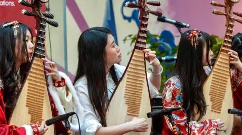 Trevor Sewell/Leicester Media Three women play large stringed, guitar like instruments, held upright