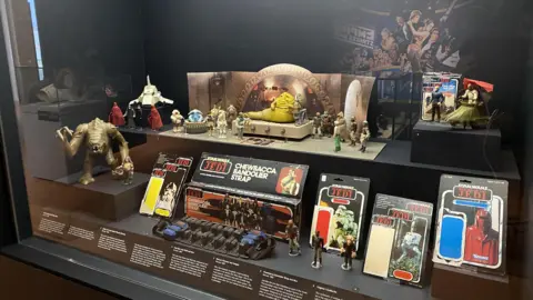 BBC A museum display behind glass of vintage Star Wars toys from 1983's Return of the Jedi film. They include orange, slug-like crime lord Jabba the Hutt who is placed on a toy throne, a brown Rancor monster with claws outstretched, and an alien musical group known as The Max Rebo Band, which features a blue elephant-like creature sitting inside a circular piano-like instrument. There are various other action figures and small vehicles on display, including opened original packaging which once contained such figures.