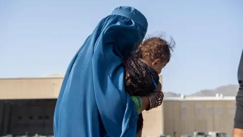 A woman dressed in blue walks outdoors at Kabul airport, cradling her young child. 