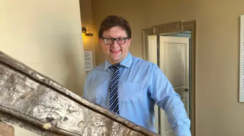 Clare Worden/BBC Andrew Stephenson stands with his hand on an old wooden staircase. He is wearing a blue shirt and striped blue tie. He has short brown hair and wears black glasses. He is smiling at the camera. 