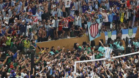 @welloffside/@simonstacpoole A large group of people, many in football shirts, are crammed behind the goal of a football pitch. A man is straddling the bar of the goalposts.