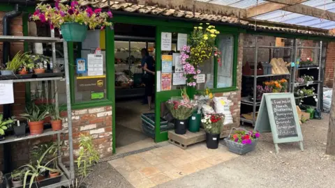 Andrew Turner/BBC Entrance to The Tacons' farm shop, a single-storey red brick building with flowers and plants outside 