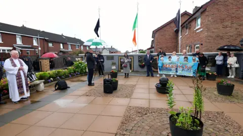Pacemaker People gathered in a paved courtyard. A priest in on the left and people in the right corner are holding up a banner reading: "Springhill-Whiterock Massacre Belfast's Bloody Sunday Time for Truth" with pictures of the five deceased.