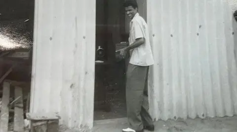 Ibraham Chisti A black and white photo of a man wearing a white shirt, dark trousers and white shoes standing in a doorway of a shack - with wooden slats on the left hand side of the photo