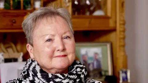 Pam Walker sitting in a chair in a living room in front of a wooden bookcase, which displays framed pictures and plants. She is wearing a black and white patterned scarf and is smiling at the camera. She has short white hair and is wearing dark pink lipstick. 