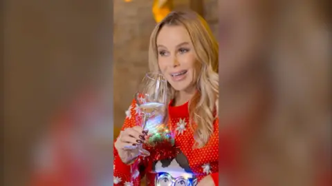 A woman, Amanda Holden, smiles while holding a wine glass. She has long blonde hair and wears a red festive jumper