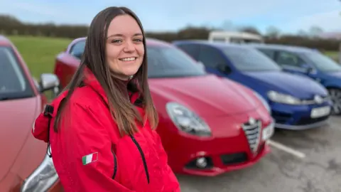 Elsie-Rose Robbins stands in front of a row of parked cars. She has long, brown hair and wears a red jacket with an Italian flag on the sleeve.