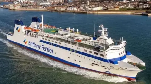 A large white and blue Brittany ferry passing close to the shore. The picture has been taken from a drone or and the top and side of the ship is visible. 