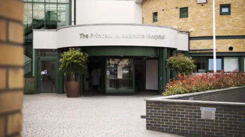 Princess Alexandra Hospital NHS Trust The entrance to the Princess Alexandra Hospital, which has green doors with silver lettering above them detailing the hospital's name. A brick path leads the way to the entrance and there are both cream and white walls beside it.
