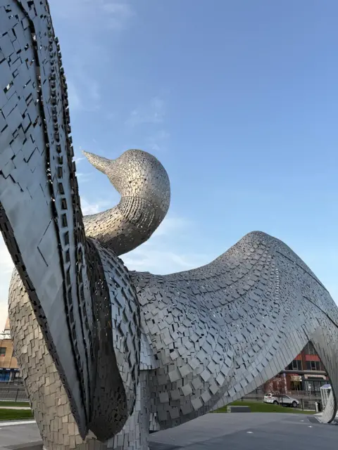 Laura Bird Rear side view of 'The Calling', large steel sculpture of a loon bird with its head thrown back and wings fully outstretched, surrounded by a paved viewing area.