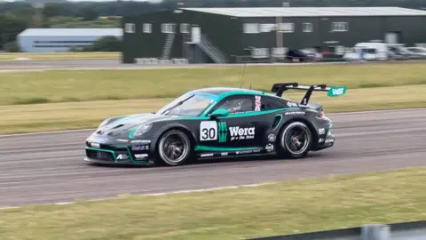 Un Porsche negro con detalles verdes alrededor de los bordes. El coche está en una pista, con césped a ambos lados y edificios al fondo. El número de carrera de Jenkins, el 30, está en el lateral del coche.