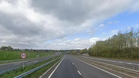 A Google Image view of the A2 near Canterbury. The road is empty and there is a sign and grass to the left, with large trees to the right.