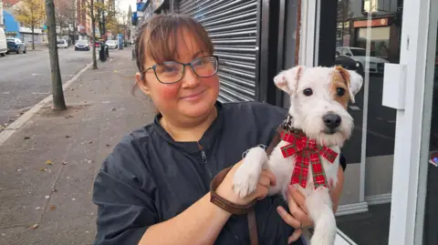 Sarah Teare. She has dark hair tied back with a fringe, wearing glasses and a black zip-up jacket. She is holding a small white dog with brown eye patch and it is wearing a Christmas bow collar. She is standing on a street footpath.