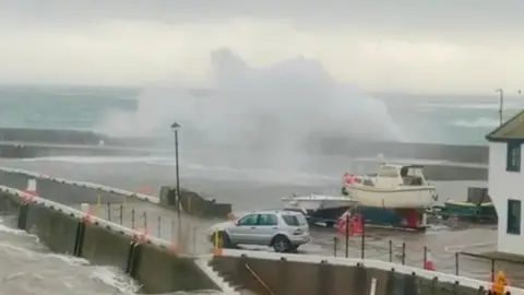 FYNODEREE DISTILLERY A wave crashes on to the harbour at Castletown, there is a boat in the foreground and a car.