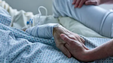 A patient in a blue spotted medical gown holding the hand of a medical practitioner