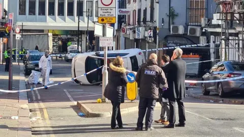 Four people, including a man with a black police jacket on, stand in front of blue and white police tape stretched across the road. Beyond the cordon is a crashed white car on its side. There is a police forensics officer in a white suit near the car.