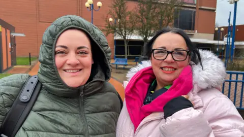 Two women wrapped up in winter clothes smile as they stand in a town centre on a rainy day outside a large red-brick building. The woman on the left is wearing a green coat, with the hood over her head. The woman on the right has black hair and is wearing a pink parka-type coat, a pink fleece and black-rimmed glasses.