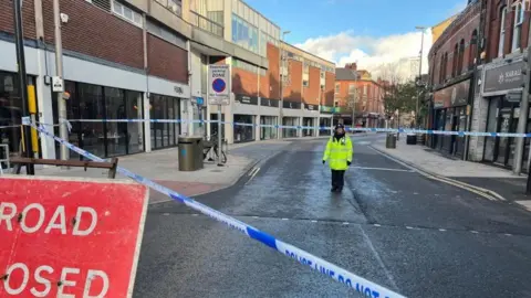 A policewoman in a yellow hi-vis jacket stands in a cordoned off area on the road with a red sign saying "road closed" outside the cordon. Shops are on both sides.