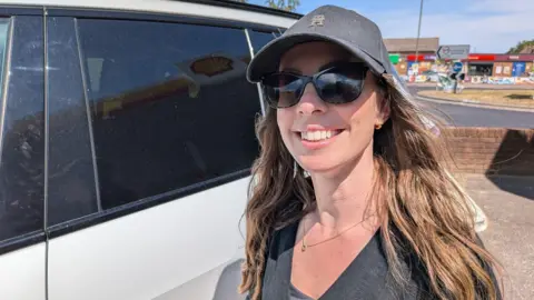 Carla Young. She has long brown hair and is wearing sunglasses and a cap. She is stood next to a white car.