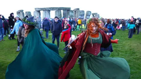 EPA Revelers dance near Stonehenge wearing green and red dresses and capes.