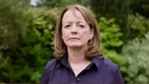 Dr Susan Gilby has mid-length hair and is wearing a purple blouse and gold necklace. She is stood in from of green bushes and is looking directly at the camera.