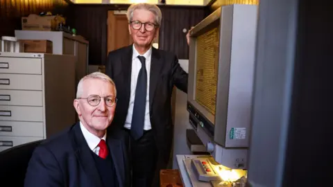 NIO A man wearing glasses and a suit is sitting looking at the camera. Another man, also wearing glasses and a suit, is standing next to him. A large grey computer with archived copy is next to them. Filing cabinets are in the background. 