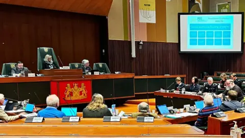 Alison Hernandez, James Vaughan and various councillors sit in a council chamber for the Police and Crime Panel.