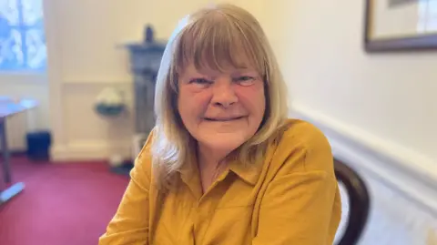 Mary Rasmussen has mid-length blonde hair with a fringe. Sitting on a chair in an office, she is wearing a dark yellow shirt.