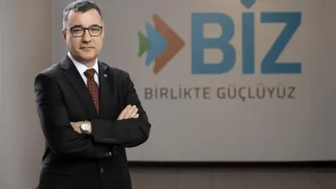 Supplied A man is wearing a suit and is standing with his arms crossed in front of a business logo. The logo says BIZ and then has some writing printed in Turkish. The man is smiling, has dark hair and is wearing glasses. 