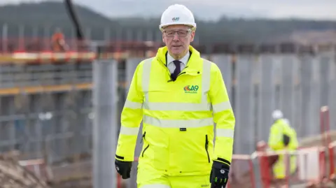 PA Media John Swinney wearing a hi-viz jacket and a white hard hat standing in front of a construction project.