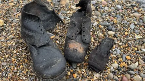Old black leather shoes, which are soaking wet and partially ruined, sat on a pebbly beach.