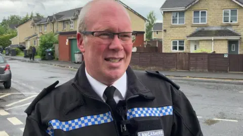 A man looks off to the right of the camera. He is wearing a black jacket with patches of white and blue checks. A patch on his chest says Lancashire. In the background, a street with several newer built houses.