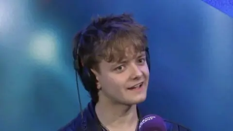 A picture of Matt Hennessy, who has mousey, short hair, sat in front of a purple microphone in a studio in front of a blue background.