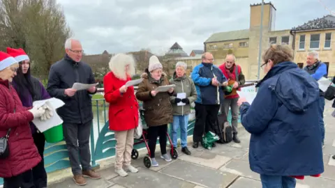 Group gathered on bridge in Chippenham read from carol sheets and singing. Two people, to the left of the image, are wearing Christmas hats. 