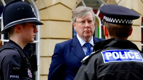 Richard Smith/BBC Roger Hirst is wearing a suit and a blue Conservative rosette while standing in a sports hall. 