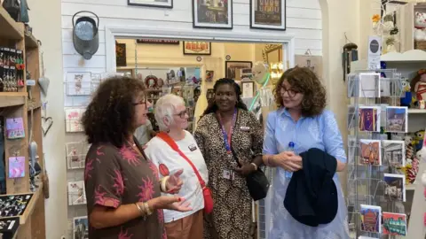 Laura Coffey/BBC Four women stood in a shop. The woman on the left is speaking to the others who are listening to her.