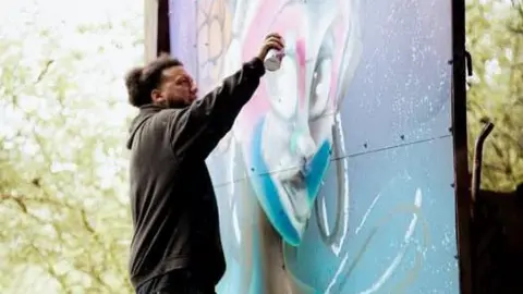 Know the unknown/Instagram A man in a black hoodie with afro hair in a bun is spraying paint onto a temporary wall. He is painting a woman's face in an animated but realistic style. There are trees behind him and the painting.