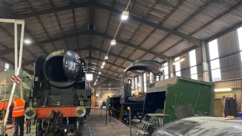 Part of a train in an SVR building and the back of a worker wearing an orange jacket. The room also has part of a locomotive, parts of trains and a set of steps visible. 