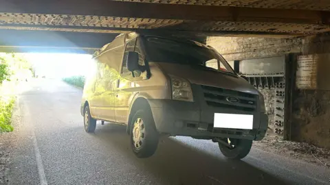 Cambridgeshire Police View of the van from under the bridge shows the roof is crumpled and dented from hitting the top of the brick structure. The front right wheel of the van has been lifted off the ground and the van is at an angle.