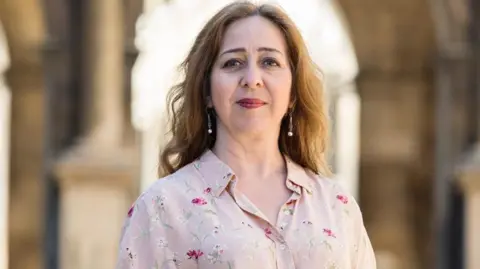 Vanessa Paloma Elbaz Vanessa Paloma Elbaz is seen long wavy brown hair wearing a floral patterned blouse, standing outdoors with a blurred architectural background.