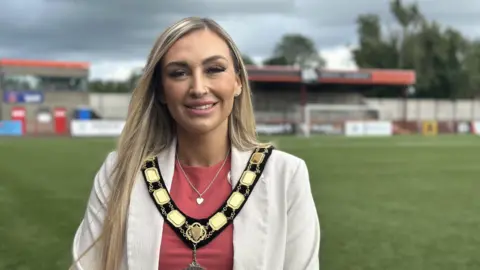 Leah Kirkpatrick has long blonde hair and wears a pink t-shirt, white jacket and a gold mayoral chain. She is standing on a football pitch.