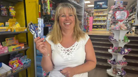 A woman with blonde hair stood smiling at the camera holding four pairs of eclipse glasses. 