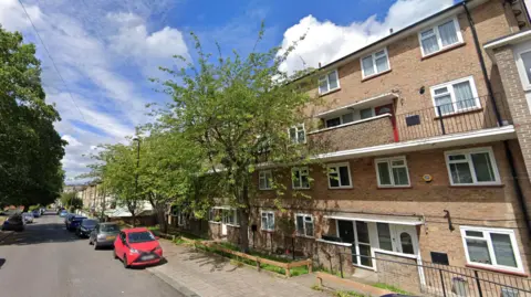 Road shows block or flats behind parked cars and trees with blue sky above