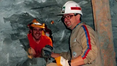Gamma-Rapho via Getty Images A man in a hard hat and beige overalls extends an arm through a hole in a rockface to clasp hands with another man in bright orange overalls. Both appear extremely happy and are smiling.