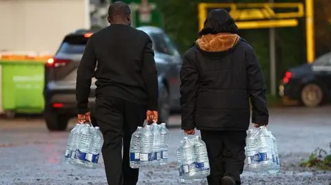 Getty Images Two people carry bottled water away from a temporary station. Their faces cannot be seen as they are walking away.