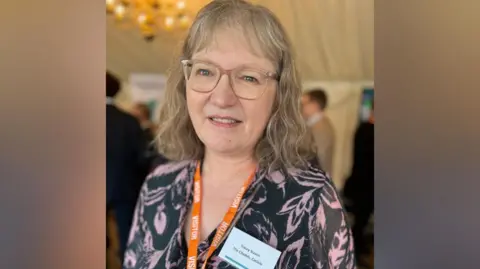 Tracey Slaven is smiling at the camera. She has short wavy blonde hair, which reaches her shoulders and a fringe. She is wearing glasses, a black top with pink floral motifs and an orange lanyard with VISITOR written on it. 