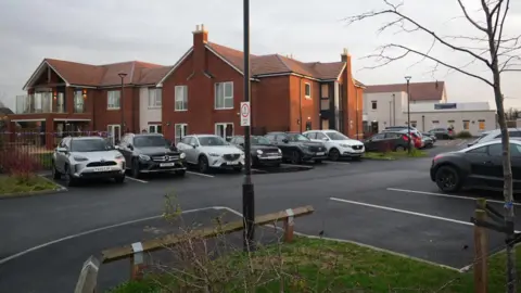 Joe Bilton / BBC A general view of the care home from the car park. There is a modern building in the background made of dark orange brick with parts painted white. There are several cars parked on a private road outside.