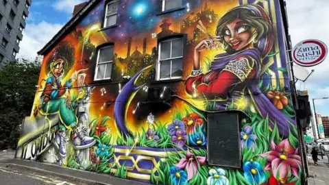 A bold, bright graphic street art wall in a sort of semi-animated style. On the left of the wall, a woman with an afro in green trousers and white trainers sits on a wall. On the right, a woman wearing long sleeved red clothing and a sari, and a purple headscarf, looks at the camera and crosses her arm. Between them is a cityscape at night with music notes floating around, and grass and flowers at the bottom.