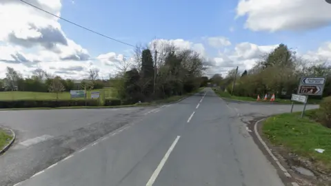 A screenshot taken from Google street view showing a junction in between several fields, with trees lining the road and a road sign pointing left to Chelworth.